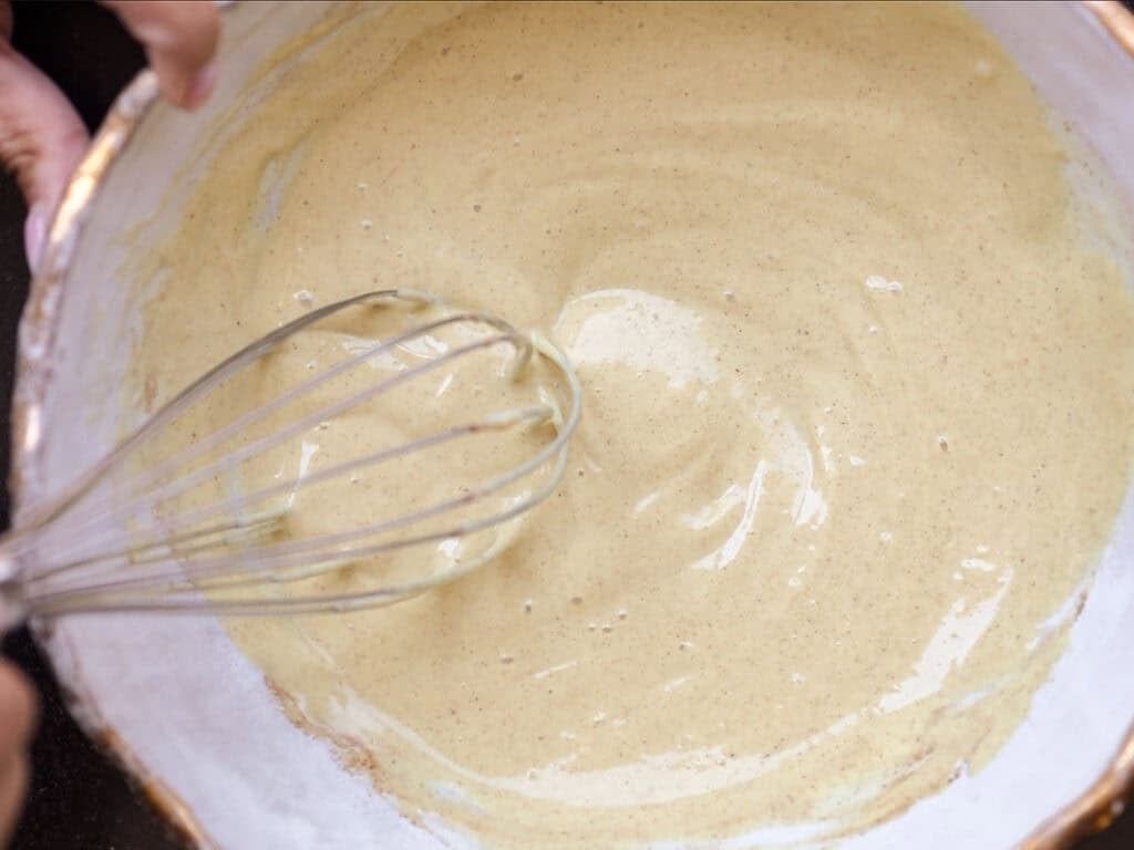 A close-up of a person whisking smooth, light yellow batter in a white bowl.